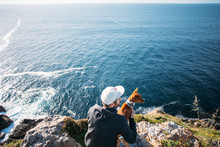 Hipster Millennial Young Man In Hoodie Sweatshirt And Five Panel Cap Sits On Cliff And Overlooks Sunset Over Ocean Together With Best Friend, Basenji Dog