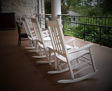 Three White Rocking Chairs Lined Up In A Balcony Of A Building Facing The Garden