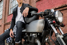 Close-up Of A Handsome Rider Biker Guy Hand In Black Leather Jacket On Helmet On Classic Style Cafe Racer Motorcycle. Bike Custom Made In Vintage Garage. Brutal Fun Urban Lifestyle. Outdoor Portrait.