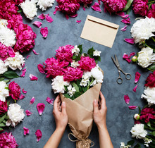 Female Florist Holding Beautiful Bouquet At Flower Shop. Florist At Work: Pretty Woman Making Summer Bouquet Of Peonies On A Working Gray Table. Kraft Paper, Scissors, Envelope For Congratulations.