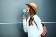 © Елена Кравчук - Positive woman licking candy, sideways, outdoor, standing, holding lollipop, hat on urban background