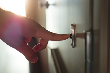 Finger pressing doorbell in sunny apartment building corridor. Close up of male hand ringing door bell in a block of flats. Salesman, fundraiser, guest or visitor behind door.