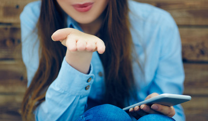 Cropped image of beautiful young girl with smart phone in hands shows air kiss gesture sitting on the background of wall of wooden boards. Photo with depth of field.