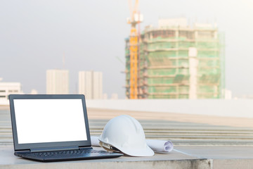 Wall Mural - The white safety helmet put on the blueprint with laptop has white screen isolated at construction site with crane background