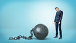 © gearstd - A small sad businessman looking down to a giant iron ball and chain on blue background.