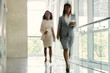© DragonImages - Cheerful businesswomen with paper cups of coffee in hands walking down office corridor with panoramic windows, shot in motion