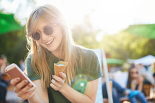 Woman Eating Icecream Using Smartphone. Portrait Of A Girl With Ice Cream Browsing Through Social Media Or Messaging Her Friends Enjoying Summer In The City Park.