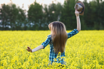 Attractive young blonde woman in blue plaid shirt straw hat enjoying her time on bright colorful blossoming yellow green meadow hard noon sun.