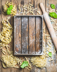 Wall Mural - Various homemade fresh uncooked Italian pasta with flour, green basil leaves, plunger and wooden tray on shabby rustic wooden background, top view, copy space, horizontal composition