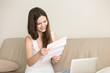 © fizkes - Smiling woman reading letter while sitting on sofa in front of laptop. Happy lady enjoying good news in written notice, pleasured with unexpected invitation to an event or celebration, loan approval