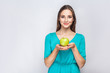 Young beautiful woman with freckles and green dress holding apple and sharing with smile. studio shot, isolated on light gray background.