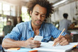 © wayhome.studio  - Candid shot of attractive bearded African college student with Afro hairstyle reading text message on his electronic device while preparing for exams at coworking space, making notes with pencil