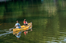 Couple Paddling In Yellow Canoe On Tree Lined Lake