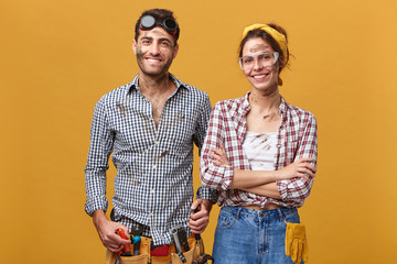 Wall Mural - Indoor portrait of positive cute young couple of professional maintenance workers wearing overalls and protective eyeglasses smiling cheerfully, enjoying helping people fixing household problems