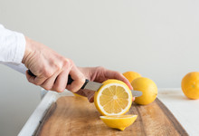 Close Up Of Middle Aged Woman's Hand Holding Knife And Slicing Lemon On Wooden Chopping Board (selective Focus)