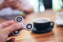 A Hand Holding And Playing Metal Silver Color Fidget Spinner With Coffee Cups And People In Background
