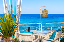 White Table And Chairs With Blue Pillows On Restaurant Terrace On Cala Nova Beach With Beautiful Sea View, Ibiza Island, Spain