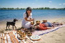 Beautiful Female Receiving Energy Sound Massage With Singing Bowls On Her Body On A River Bank At Spring Sunny Day. And Little Black Doggy Interested In