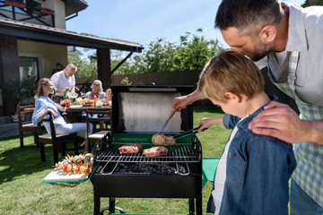 Wall Mural - Close-up view of father and son grilling meat while family sitting at table outdoors