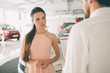 © estradaanton - friendly car salesman talking to a young woman and showing a new car inside showroom Signing of contract.