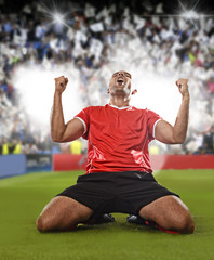 happy and excited football player in red jersey celebrating scoring goal kneeling on grass pitch