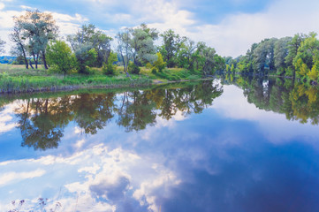 Calm beautiful scenic landscape with blue river, green trees and reflecting in water with cloudy sky. Magical sunset over the river in rural terrain. Natural, wild landscape.