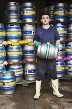 Man Working In A Brewery, Standing Next To A Stack Of Metal Beer Kegs, Holding Beer Keg.