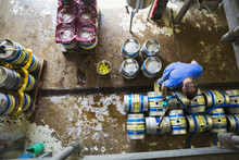 Directly Above View Of A Man Working In A Brewery, Metal Beer Kegs Standing On The Floor.