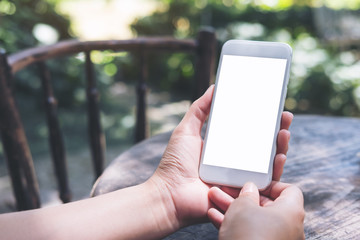 Mockup image of hands holding white mobile phone with blank screen on vintage wooden table and nature background