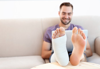 Handsome young man with laptop and bandaged leg sitting on sofa at home, closeup