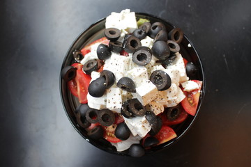 Greek salad with sirtaki cheese and black olives and a red tomato. On white cheese is ground pepper. Salad in a glass plate is on a dark table.