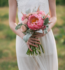 Wall Mural - bride in a dress standing in a green garden and holding a wedding bouquet of flowers and greenery. Woman holding colorful bouquet with her hands on wedding day