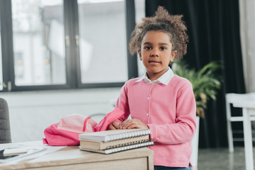 Wall Mural - Adorable little girl standing with backpack and looking at camera indoors