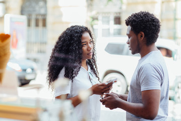 Wall Mural - Young african american couple with smart phones in hands are facing each other in european street of city center.