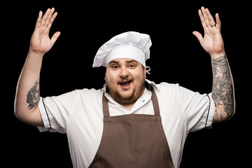 happy young chef in hat and apron gesturing with hands isolated on black