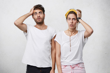 Wall Mural - Human facial expressions, emotions, feelings, reaction and attitude. Indoor shot of shocked forgetful young Caucasian man and woman holding hands on heads and looking at camera with shock and terror