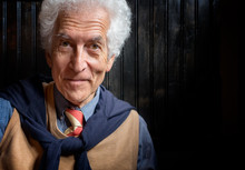 Senior Man Portrait Looking At Camera. Kind Face. Wearing Tie And Sweater Tied Over The Shoulders. White Curly Hair And Bright Intelligent Eyes. Close Up With Copy Space.