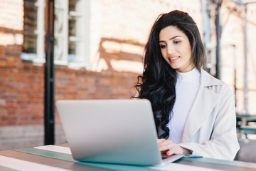 Adorable brunette woman in white clothes sitting outdoors at cafe using her laptop computer communicating with friends online using free internet connection. People, lifestyle, technology concept