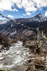 Black mountain with snow on the top. On the ground is frozen river with stone, ice, yellow grass and Tibetan prayer flags at Thangu and Chopta valley in winter in Lachen. North Sikkim, India.