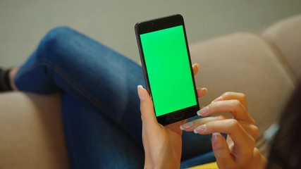 Close up woman using smartphone with green screen while sitting on the couch in the livinng room. Woman hands scrolling, typing, tapping on touchscreen. chroma key. top view