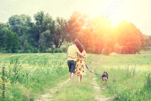 Happy couple in love with dog walking on a rural dirt road in springtime at sunset. The woman and man hugging. The woman keeps her dog on a leash. Couple and dog back to the camera. Casual style.
