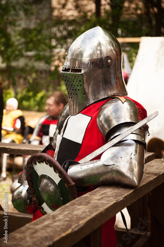 Plakat Kostium z epoki średniowiecza w turnieju rycerskim. Średniowieczna rekonstrukcja historyczna - mężczyzna w metalowym hełmie i czerwonym zbroi, trzymający miecz i tarczę