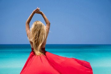 Beautiful young woman with blond long hair in red fluttering dress is standing a back on the coastline of azure caribbean sea