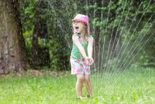 Happy Toddler Girl Playing In A Sprinkler On A Hot Summer Day