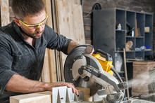 Young Brunette Man In Black Overalls By Profession Carpenter Builder Saws With A Circular Saw A Wooden Board On A Wooden Table In The Workshop
