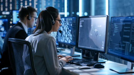 Aufkleber - Female working in a Surveillance Team Gives Instructions with the Help of the Headsets. In the Background People Working and Monitors Show Various Information.