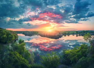 Wall Mural - Amazing scene with river, green trees, rocks and amazing blue sky with colorful clouds reflected in water at sunset. Fantastic summer landscape with lake, overcast sky and yellow sun in the evening
