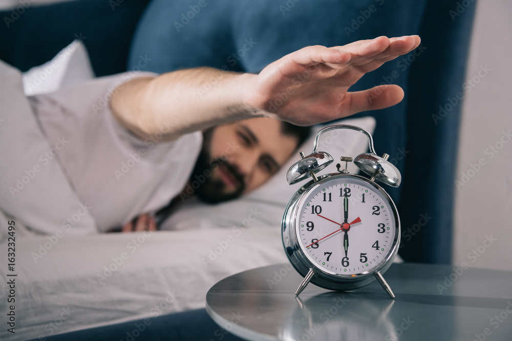 Bearded young man trying to turn off alarm clock while lying in bed at morning - obrazy, fototapety, plakaty 