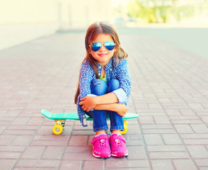 Wall Mural - Fashion happy smiling little girl child sitting on the skateboard in the city