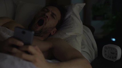 Young adult man can't sleep, yawning and browsing the net on cellphone, insomnia